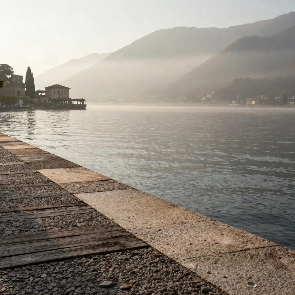 Riva di pietra del Lago di Como con ville lontane, acqua immobile e montagne velate al mattino